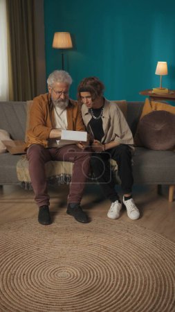 Photo for Shot of a teenager, young man sitting with his grandfather on a sofa, couch, reading a document, paper, application response, letter. College, university, enrollment, family values. - Royalty Free Image