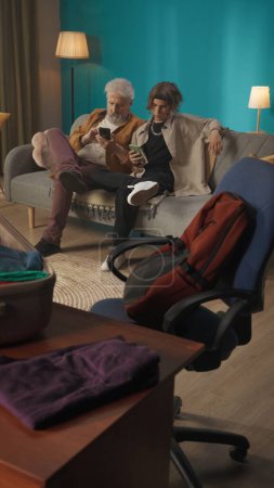 Foto de Foto de un adolescente, joven con su abuelo, abuelo sentado en un sofá, sofá y utilizando sus teléfonos inteligentes, desplazamiento, mensajes de texto. Abre la maleta en primer plano. Mudarse a un nuevo lugar, familia. - Imagen libre de derechos