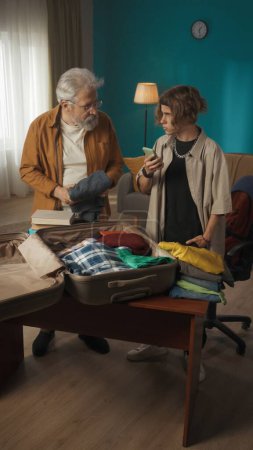 Photo for Shot of a teenager, young man packing his belongings in a suitcase. His grandfather, granddad is helping him with that. The boy is holding a smartphone. College, university, moving to a new place. - Royalty Free Image