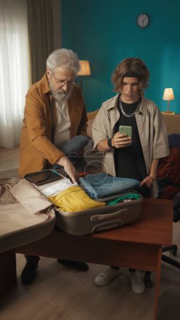 Photo for Shot of a teenager, young man packing his belongings in a suitcase. His grandfather, granddad is helping him with that. The boy looks at his phone. College, university, moving to a new place. - Royalty Free Image