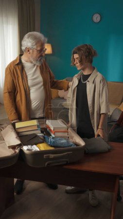 Photo for Shot of a teenager, young man packing his belongings in a suitcase. His grandfather, granddad is helping him with that. The boy looks frustrated. College, university, moving to a new place. - Royalty Free Image