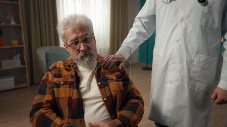 Photo for Medium shot of a handicapped eldery patient on a wheelchair having a medical check up at home. The doctor is patting him on the shoulder. Healthcare system, medical service, advertisement. - Royalty Free Image