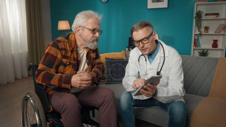 Photo for Full-size shot of a doctor checking up the condition of his patient at home. He is talking to an eldery man on a wheelchair, showing something on a tablet. Healthcare, medical service, advertisement. - Royalty Free Image