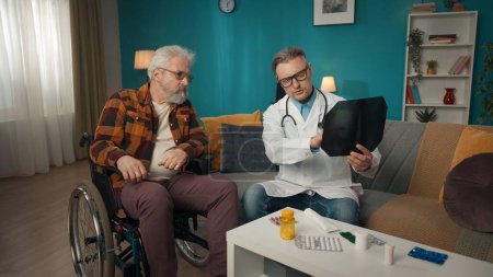 Photo for Full-size shot of a doctor checking up the condition of his patient at home. He is talking to an eldery man on a wheelchair, showing a x-ray picture, speaking. Healthcare, medical, advertisement. - Royalty Free Image