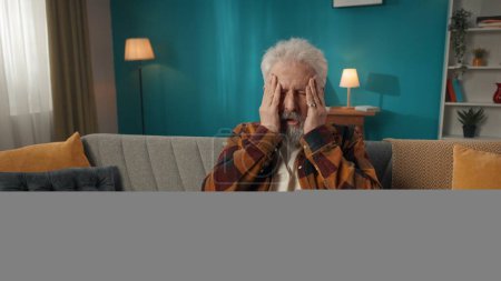 Téléchargez les photos : Plan moyen d'un vieillard retraité assis sur un canapé, massant ses tempes et sa tête à cause d'un grave mal de tête, migrane. Système de soins de santé, service médical, publicité. - en image libre de droit