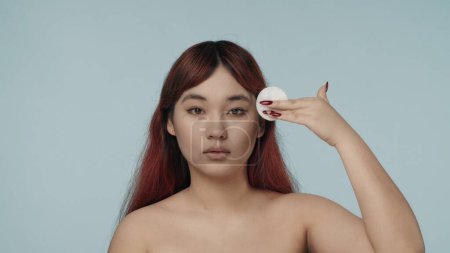 Photo for Close-up isolated photo of a young seminude woman with red dyed hair and nude makeup looking at the camera, moving a cotton pad across her face and body. Beauty, skincare advertisement. - Royalty Free Image