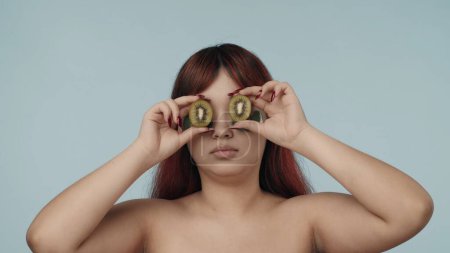 Photo for Close-up isolated photo of a young seminude woman with red dyed hair and nude makeup wearing eyepatches, holding two kiwis in front of her eyes. Beauty, cosmetics, skincare routine advertisement. - Royalty Free Image