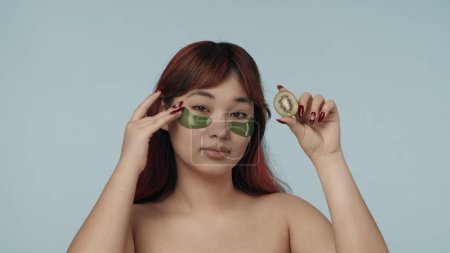 Photo for Close-up isolated photo of a young seminude woman with red dyed hair and nude makeup wearing eyepatches, touching them gently, holding a cut kiwi. Beauty, cosmetics, skincare routine advertisement. - Royalty Free Image