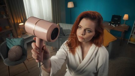 Photo for Close-up shot from above of a young girl with medium-length red hair and makeup standing in a bathrobe holding a hair dryer above her and looking at it skeptically, prejudiced with uncertainty. - Royalty Free Image