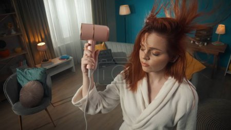 Photo for In a close-up shot from above stands a young girl with red hair and makeup in a bathrobe holding a hair dryer above her. She is drying her hair with her eyes closed. Hair from the air flow develops. - Royalty Free Image