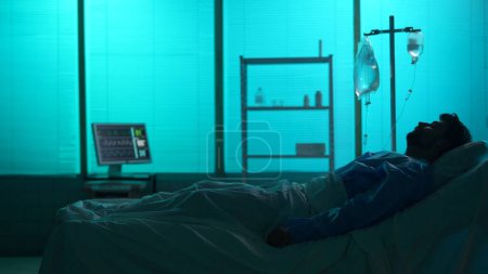Foto de Foto de silueta de tamaño completo de un hombre enfermo acostado o durmiendo en una cama de hospital conectada a una cámara de goteo. Unidad de cuidados intensivos, monitor de pie en un segundo plano. Hospital, tratamiento impaciente - Imagen libre de derechos