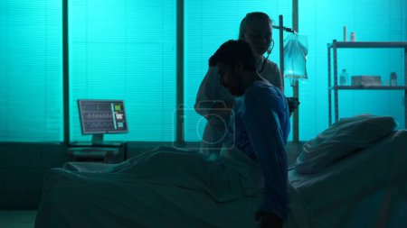 Foto de Una silueta de tamaño completo de un paciente enfermo acostado en una cama de hospital. Una doctora, la enfermera le hace un chequeo, escuchando sus pulmones con estetoscopio. Hospital, tratamiento impaciente, médico. - Imagen libre de derechos