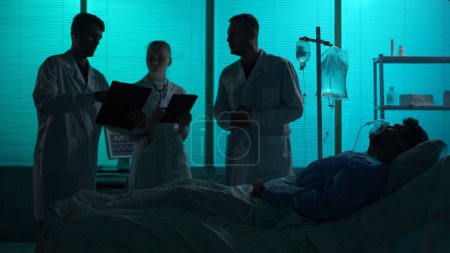 Foto de Foto de silueta de tamaño completo de tres médicos parados alrededor de un paciente en un soporte respiratorio, en coma, acostados en una cama de hospital frente a ellos. Hospital, tratamiento impaciente, atención médica. - Imagen libre de derechos