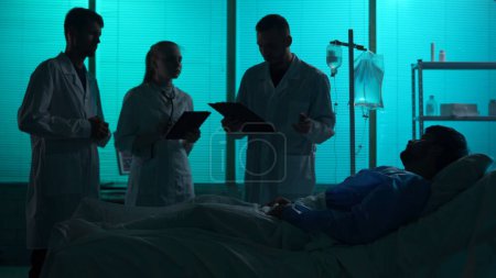 Photo for Full-sized silhouette shot of three doctors standing around a patient laying in a hospital bed in front of them. Doctors discuss the treatment method with him. Hospital, impatient treatment, medical. - Royalty Free Image
