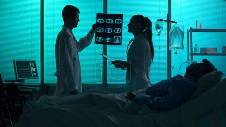 Téléchargez les photos : Silhouette pleine grandeur de deux médecins debout derrière un patient sur un support respiratoire, dans le coma. Les médecins regardent ses poumons, discutent de la méthode de traitement. Hôpital, soins médicaux. - en image libre de droit