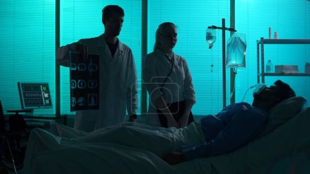 Foto de Foto de silueta de dos doctores parados junto a un paciente acostado en una cama de hospital. Los médicos muestran su ct pulmonar y discuten el método de tratamiento con él. Hospital, impaciente, médico. - Imagen libre de derechos