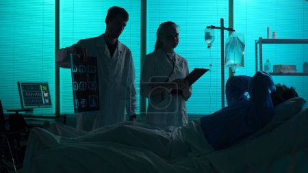 Foto de Foto de silueta de dos doctores parados junto a un paciente acostado en una cama de hospital. Los doctores muestran su ct pulmonar. El paciente se agarra la cabeza en la desesperación y la negación. Hospital, atención médica. - Imagen libre de derechos