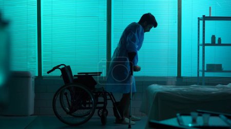 Photo for Full-sized silhouette shot of a disabled man, patient with mobility impairment trying to get up from a wheelchair, making a few succesful steps leaning on a crutch. Hospital, rehabilitation, medical. - Royalty Free Image