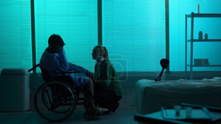 Téléchargez les photos : Silhouette pleine grandeur d'un homme handicapé, patient à mobilité réduite en fauteuil roulant, pleurant dans le désespoir et le déni à côté de son bien-aimé, jeune femme. Hôpital, réadaptation, médical. - en image libre de droit