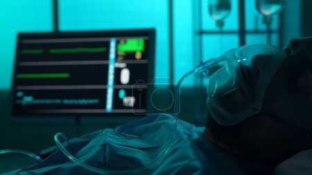 Photo for Close-up detail shot of a patient on breathing support dying in a hospital bed. His ICU monitor on the background displays loss of heart and brain activity. Hospital, emergency, resuscitation. - Royalty Free Image