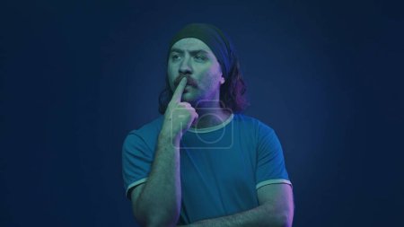 Téléchargez les photos : Portrait d'un homme moustachu portant un bandeau dans un studio sur fond bleu en néon rose et vert. Un homme réfléchit intensément à quelque chose, en mettant son doigt sur ses lèvres - en image libre de droit