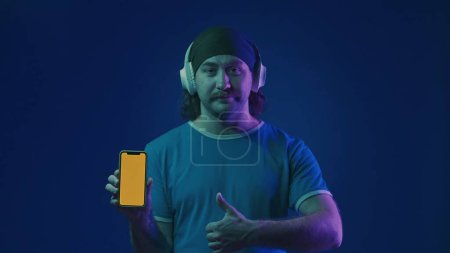 Téléchargez les photos : Un homme en casque sans fil blanc tient un téléphone avec un écran vert et montre un pouce vers le haut. Un homme en studio sur fond bleu en néon rose et vert - en image libre de droit