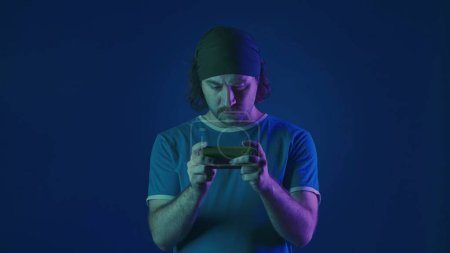Téléchargez les photos : Un homme tendu joue à un jeu en ligne sur son smartphone. Un homme dans un studio sur fond bleu en néon rose et vert - en image libre de droit