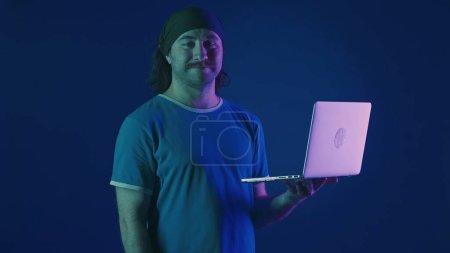 Foto de Un hombre usa un portátil para trabajar. Un hombre con un portátil en la mano en un estudio sobre un fondo azul en luz de neón rosa y verde. Trabajo remoto, freelancer - Imagen libre de derechos