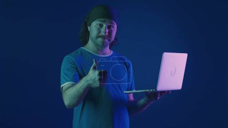 Foto de Un hombre usa un portátil para trabajar y muestra los pulgares hacia arriba. Un hombre con un portátil en la mano en un estudio sobre un fondo azul en luz de neón rosa y verde. Trabajo remoto, freelancer - Imagen libre de derechos