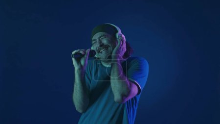 Photo for A mustachioed man in white wireless headphones dances and sings into a microphone. Woman in studio on blue background with pink and green neon lights. Karaoke - Royalty Free Image