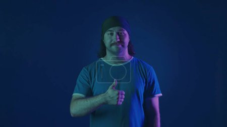 Foto de El hombre muestra los pulgares arriba. Retrato de un hombre de pelo largo bigotudo en un estudio sobre un fondo azul en luz de neón rosa y verde - Imagen libre de derechos