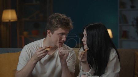Photo for First relationships creative concept. Portrait of teenage couple spending leisure time.. Boy and girl sitting on the sofa and eating pizza, shyly looking at each other. - Royalty Free Image