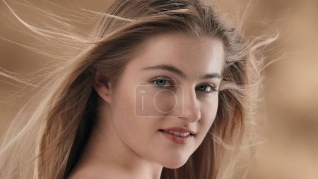 Photo for A close up of a young blonde-haired woman against a stained background. With her head slightly turned and tilted, she looks at the camera and smiles. The wind is blowing on her, developing her hair. - Royalty Free Image