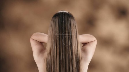 Téléchargez les photos : Dans le cadre, une jeune femme aux cheveux longs, raides et blonds se dresse sur un fond tacheté. Elle se tient le dos à la caméra, fixant ses cheveux des deux mains. Tir moyen. - en image libre de droit
