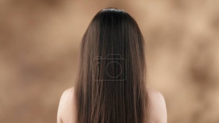 Foto de En el marco contra un fondo manchado es una mujer de mediana edad con el pelo largo, oscuro y liso. De pie con su espalda a la cámara demuestra la belleza y la salud de su cabello. - Imagen libre de derechos