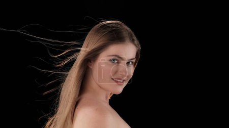 Photo for A young woman without makeup on a dark background. She stands sideways and looks at the camera and smiles, the light falls on her and the wind blows. It blows her hair in different directions. - Royalty Free Image