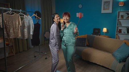 Photo for Full-sized photo capturing two young women taking photos of themselves on a smartphone using a selfie stick in a room. Sparkly costumes hanging on a background. Sleepover, friendship, siblings. - Royalty Free Image