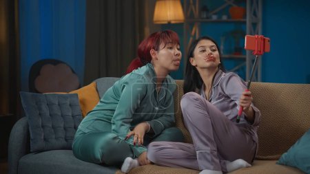 Photo for Medium-full photo capturing two young women wearing pajamas sitting on the couch and doing their evening skincare routine, taking selfies, laughing. Girls night, sleepover, friendship, siblings. - Royalty Free Image