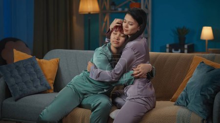 Photo for Medium-full photo capturing two young women wearing pajamas sitting on the couch in a room, hugging. They made up after a fight. Girls night, sleepover, friendship, siblings, supporting. - Royalty Free Image