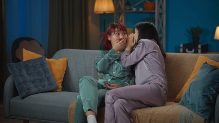 Photo for Medium-full photo capturing two young women wearing pajamas sitting on the couch in a room. One of them shares a gossip, secret to her friend, sister. Girls night, sleepover, friendship, siblings. - Royalty Free Image