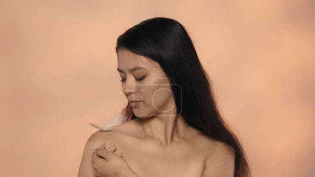 Photo for Beauty and skincare treatment creative concept. Closeup studio shot of attractive young model. Beautiful asian girl with long hair and natural makeup holding white small feather on her shoulder. - Royalty Free Image