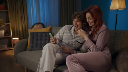 Photo for Beauty and healthy relationships advertisement concept. Portrait of young couple spending time together. Man and woman in pajamas sitting on the sofa, holding smartphone laughing. - Royalty Free Image