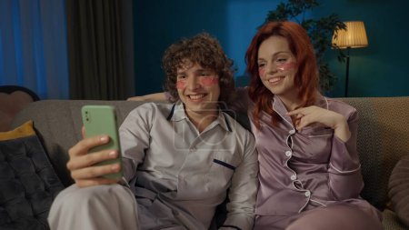 Photo for Beauty and healthy relationships advertisement concept. Portrait of young couple spending time together. Man and woman sitting on the sofa, wearing pajamas and eyepatches, taking selfie photos. - Royalty Free Image