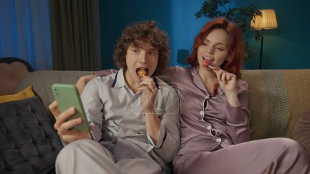 Photo for Beauty and healthy relationships advertisement concept. Portrait of young couple. Closeup of man and woman in pajamas sitting on the sofa with lollipops, watching something on smartphone. - Royalty Free Image