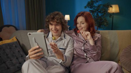 Photo for Beauty and healthy relationships advertisement concept. Portrait of young couple. Closeup of man and woman in pajamas sitting on the sofa with lollipops, watching something on tablet. - Royalty Free Image
