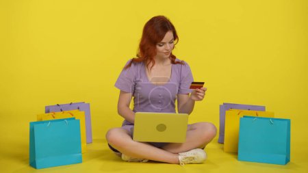 Foto de Una mujer hace una compra en una tienda en línea utilizando un ordenador portátil y una tarjeta de crédito. Mujer pelirroja se sienta con las piernas cruzadas en el suelo, con un ordenador portátil en su regazo, rodeado de bolsas de compras en el estudio en un - Imagen libre de derechos