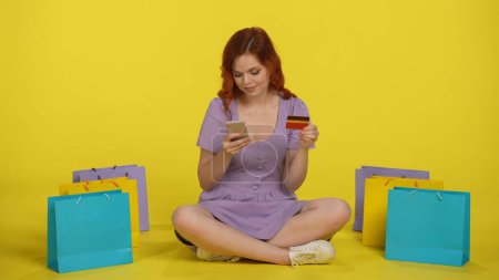 Foto de Una mujer hace una compra en una tienda en línea con un teléfono inteligente y una tarjeta de crédito. Mujer pelirroja se sienta con las piernas cruzadas en el suelo rodeado de bolsas de compras en el estudio sobre un fondo amarillo. - Imagen libre de derechos