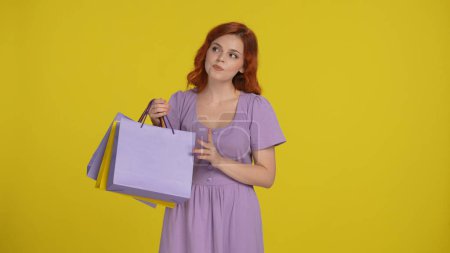 Photo for A young woman is thinking intently, tapping her fingers on shopping bags. Woman in the studio on a yellow background - Royalty Free Image