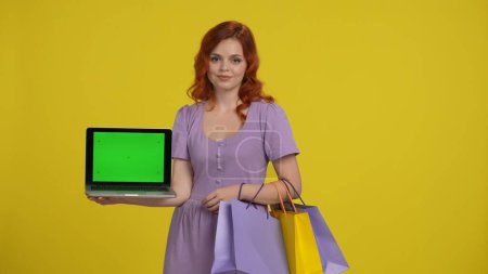 Téléchargez les photos : Femme rousse tient un ordinateur portable avec un écran vert. Femme avec sacs à provisions en studio sur fond jaune. Espace publicitaire, espace de travail. Vente, Black Friday, réductions - en image libre de droit