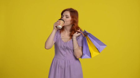 Téléchargez les photos : Une femme rousse tient des sacs à provisions et boit du café, du thé dans une tasse en papier. Jeune femme en vêtements d'été isolé sur fond jaune. Concept de vente de rabais sur les achats. Modélisation d'espace de copie - en image libre de droit